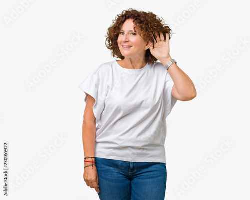 Beautiful middle ager senior woman wearing white t-shirt over isolated background smiling with hand over ear listening an hearing to rumor or gossip. Deafness concept.