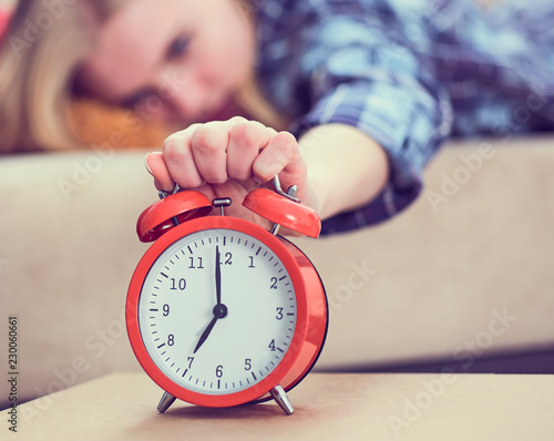 Young girl lies on the couch and stretches her hand to the red alarm clock to turn it off. Late wake up.