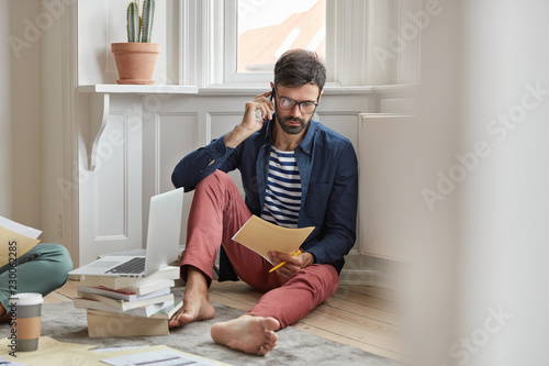 People and business concept. Unshaven male employee thinks on better solution, talks via smart phone, reads documentation, sits bare foot, solves problems, discuss working process, plan organization