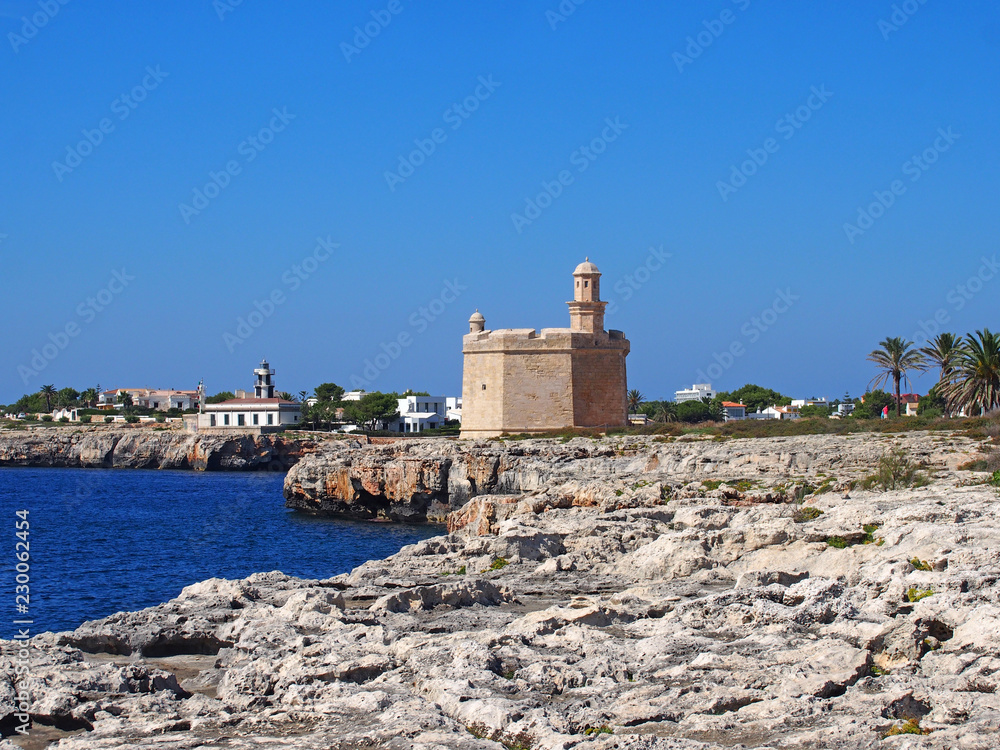 a bright sunlit view of the cliffs and coast in ciutadella menorca with deep blue sea and rocky cliffs with the historic castle and lighthouse in the distance