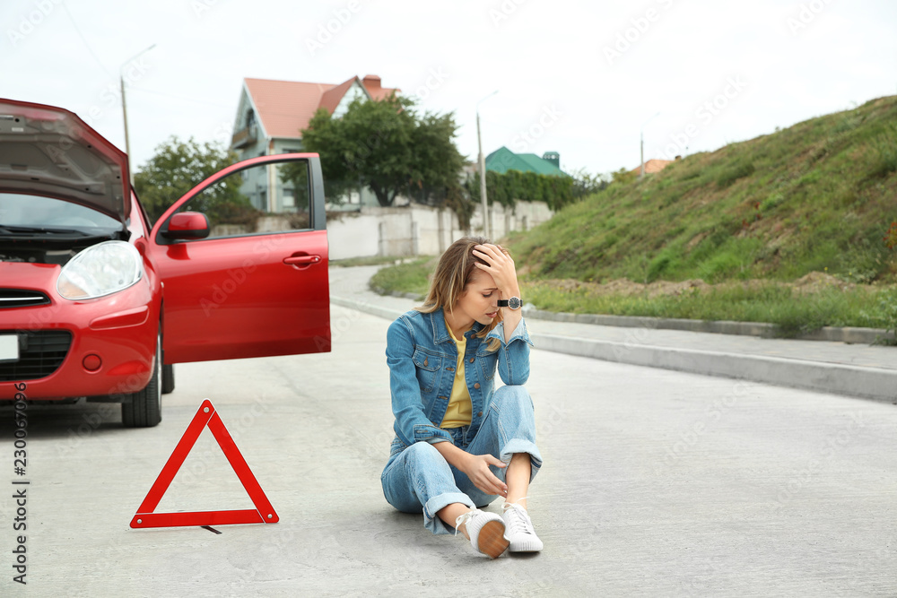 Upset woman sitting near warning triangle and broken car on road