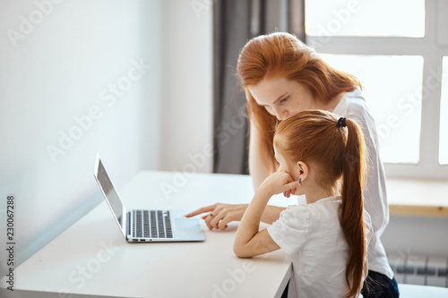 Loving caucasian red-haired mummy and her, sweetheart girl sitting at table and looking at the laptop, watching new TV chanal for kids. photo