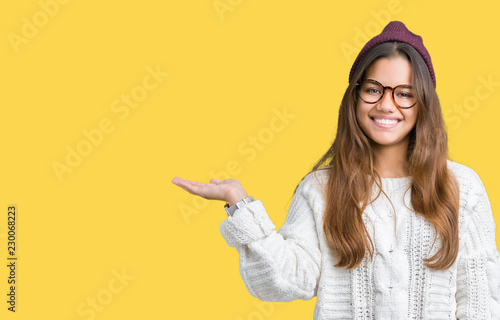 Young beautiful brunette hipster woman wearing glasses and winter hat over isolated background smiling cheerful presenting and pointing with palm of hand looking at the camera.