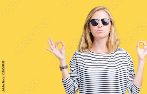Beautiful young woman wearing sunglasses over isolated background relax and smiling with eyes closed doing meditation gesture with fingers. Yoga concept.