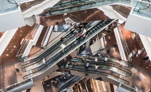 Customers clients moving on escalator staircases inside of giant modern shopping mall. Consumption concept. photo