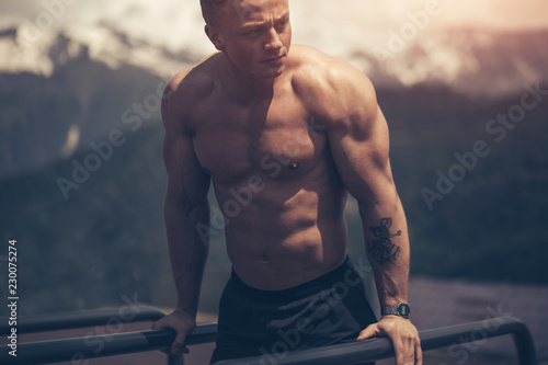 Motivated athletic shirtless man does bodyweight push ups on low horizontal bars at cool mountains area outdoor gym in the early morning. photo