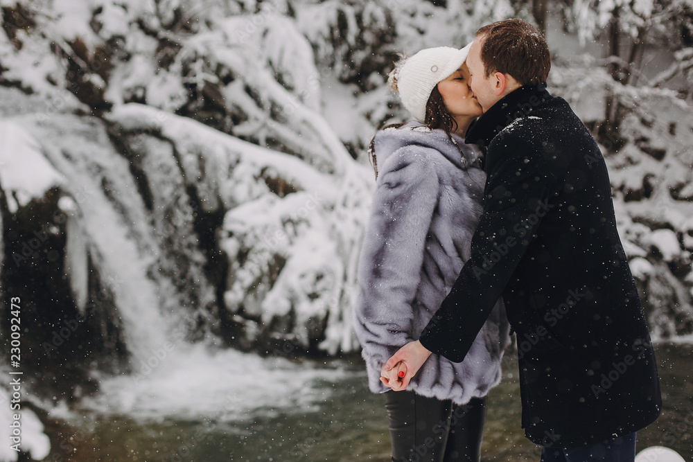 Stylish couple kissing in winter snowy mountains. Happy romantic man and woman in luxury clothes gently embracing at waterfall in snow. Holiday getaway together. Space for text
