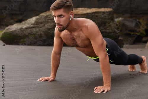 People, endurance and motivation concept. Concenrated fitness young man instructor does plank exercise, prepares for sport competitions, does sports outdoor, listens music for entertainment.