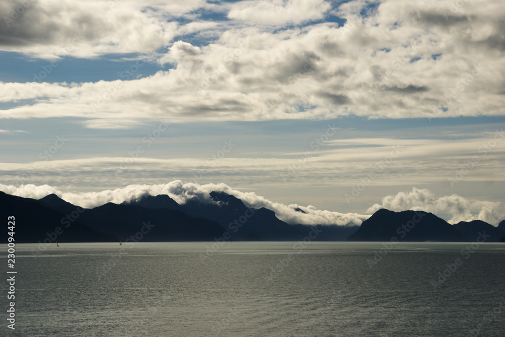 Mountains with clouds laying on mountain top under sunny blue sky with clouds looking across the ocean in Seward Alaska