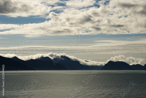 Mountains with clouds laying on mountain top under sunny blue sky with clouds looking across the ocean in Seward Alaska