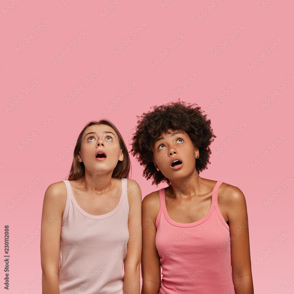 Vertical shot of scared fearful mixed race women stare with bugged eyes and widely opened mouthes upwards, dressed in casual vest, see something unbelievable. Omg, what is going on upstairs? Vertical shot of scared fearful mixed race women stare with bugged eyes and widely opened mouthes upwards, dressed in casual vest, see something unbelievable. Omg, what is going on upstairs?