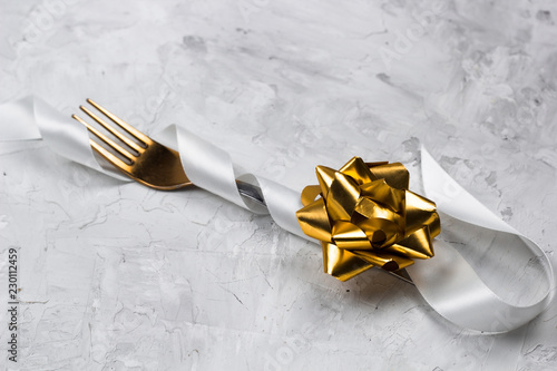 Festive table decoration for a dinner, golden fork with white shiny satin ribbon and golden bow, concrete stone background