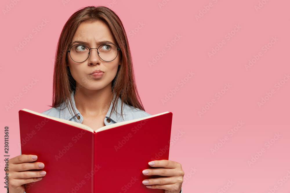 Uncertain brunette woman purses lips and has doubtful facial expression, thinks about future examination, ponders on further developing of plot in romantic story, carries book in front, likes reading Uncertain brunette woman purses lips and has doubtful facial expression, thinks about future examination, ponders on further developing of plot in romantic story, carries book in front, likes reading