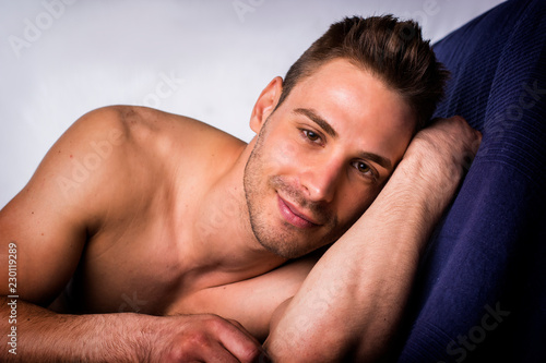 Cute, happy expression, naked young smiling man on blue sheets looking at camera, resting on his arm