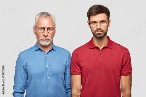 Father and son have serious expressions, stand closely against white background, have good relationships. Mature handsome man in formal shirt and spectacles stands near his young grandson indoor