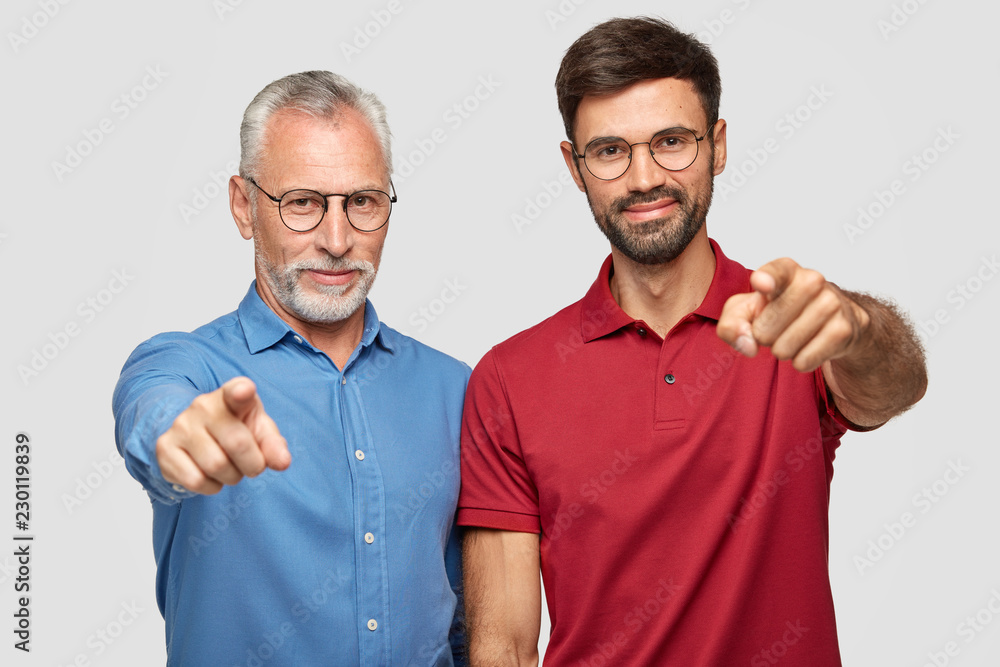Studio shot of elderly man and bearded male adult stand next to each other indoor, point directly at camera, express their choice, indicate at something in front, have pleased facial expressions Studio shot of elderly man and bearded male adult stand next to each other indoor, point directly at camera, express their choice, indicate at something in front, have pleased facial expressions