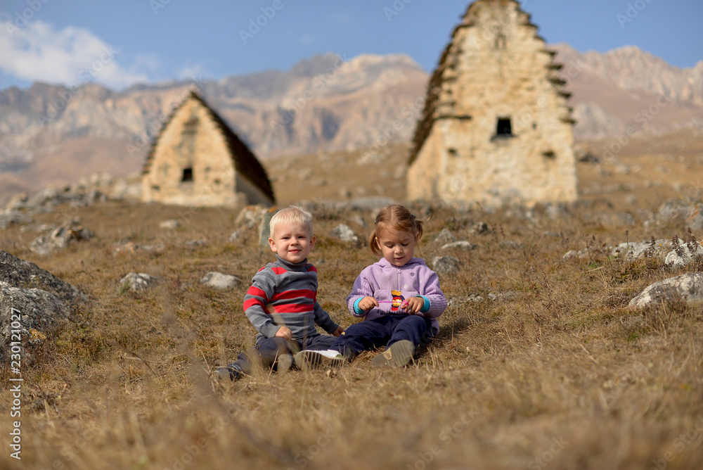 Young children sit in the dry grass on the background of mountains and ancient Ossetian buildings in North Ossetia - Alania. Young children sit in the dry grass on the background of mountains and ancient Ossetian buildings in North Ossetia - Alania.
