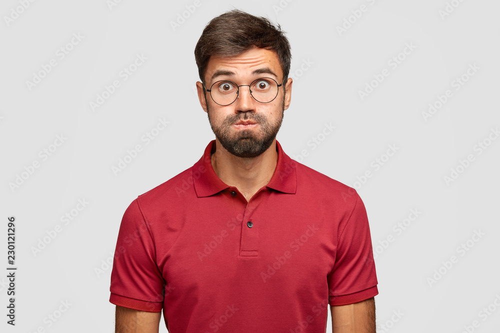 Studio shot of emotive man has displeased expression, blows cheeks, has eyes popped out, wears round spectacles, casual clothes, models against white background. Young man grimaces, requires attention Studio shot of emotive man has displeased expression, blows cheeks, has eyes popped out, wears round spectacles, casual clothes, models against white background. Young man grimaces, requires attention