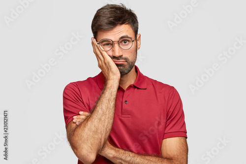 Принти на полотні Indoor shot of sad man looks bored, keeps hand on cheek, dressed in red casual clothes, feels lonliness, doesnt know what to do, poses against whitte studio wall