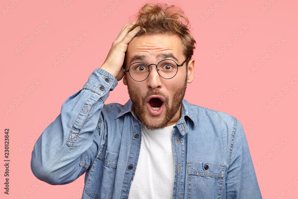 Shocked young European man keeps hand on head, jaw dropped, hears terrible things from friend, wears glasses for good vision, expresses surprisement, isolated over pink studio wall. Omg concept Shocked young European man keeps hand on head, jaw dropped, hears terrible things from friend, wears glasses for good vision, expresses surprisement, isolated over pink studio wall. Omg concept