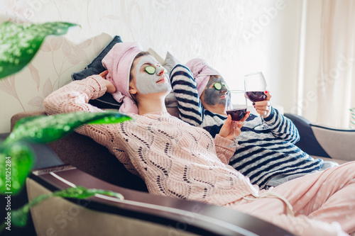 Mother and her adult daughter applied facial masks and cucumbers on eyes. Women chilling while having wine