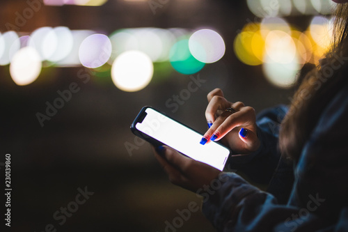 Woman pointing finger on blank screen smartphone on background bokeh light in night atmospheric city. hipster using in hands clean gadget mobile phone closeup, mockup street, online wifi internet