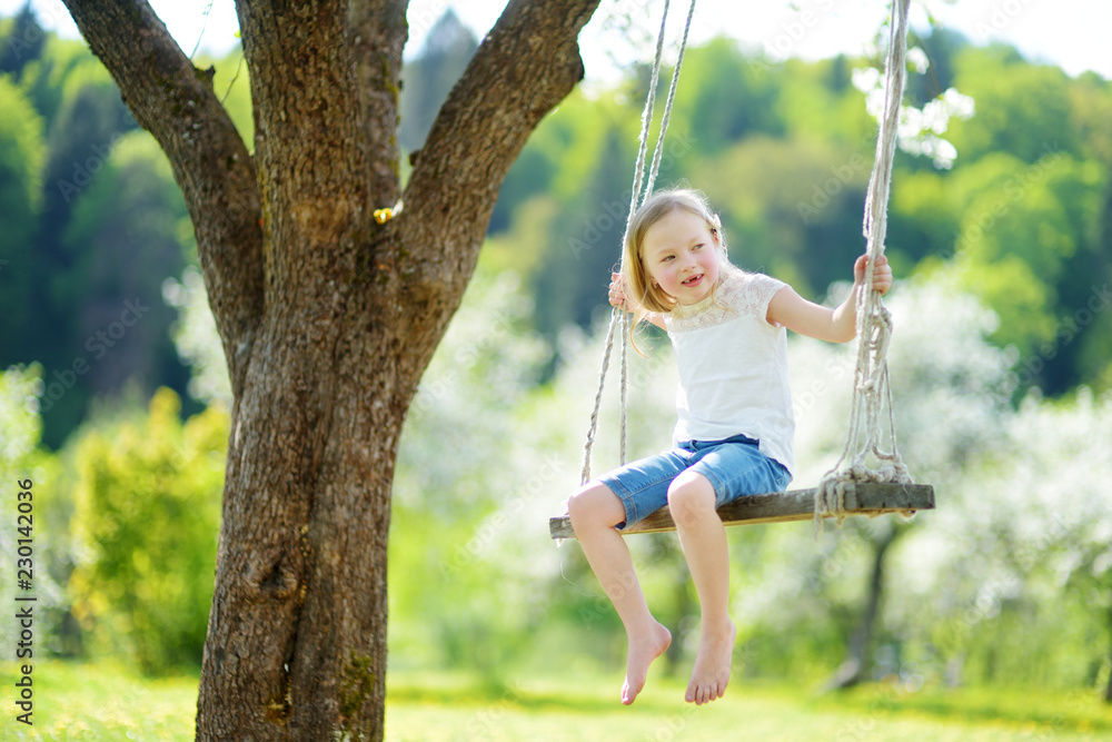 Cute little girl having fun on a swing in blossoming old apple tree garden outdoors on sunny spring day.