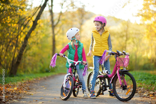 Cute sisters riding bikes in a city park on sunny autumn day. Active family leisure with kids. Children wearing safety hemet while riding a bicycle.