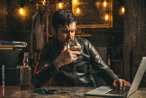 Bad news in messenger. Drunk man in a vintage pub used laptop. Bearded Man drinks brandy or whiskey. Serious man having alcohol addiction. Social concept.