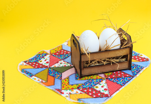 White eggs in a wooden crate, on a patchwork rug, with yellow background.