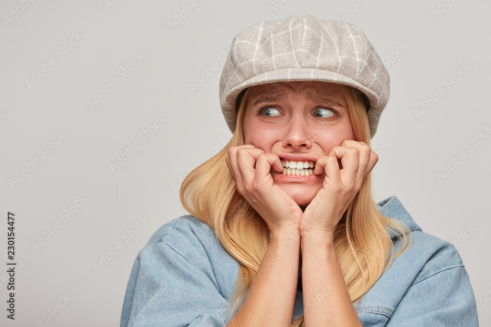Scared blonde girl looks aside frightened afraid chattering teeth with fear,bites nails from fright, hands holding cheeks, on grey background Scared blonde girl looks aside frightened afraid chattering teeth with fear,bites nails from fright, hands holding cheeks, on grey background