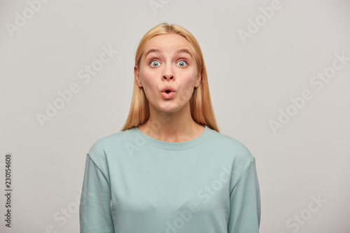 Emotional blonde young woman, looking very surprised, open-eyed, whistles in surprise, wearing blue casual sweatshirt, see something unexpected in front. Human reaction concept over grey background photo