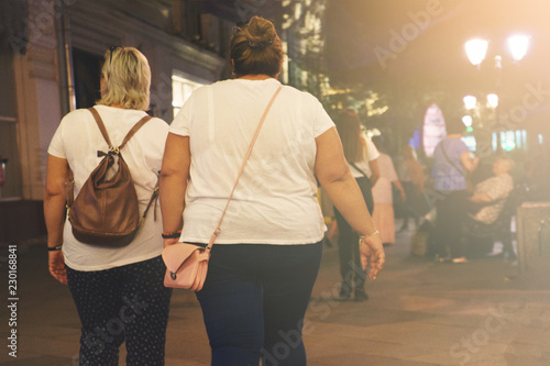 Two fat women with a backpack and bag go after shopping in the evening street of the city.
