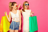 Portrait of two young stylish smiling blond women holding shopping bags. Girls dressed in summer hipster clothes. Positive models posing over pink blackground