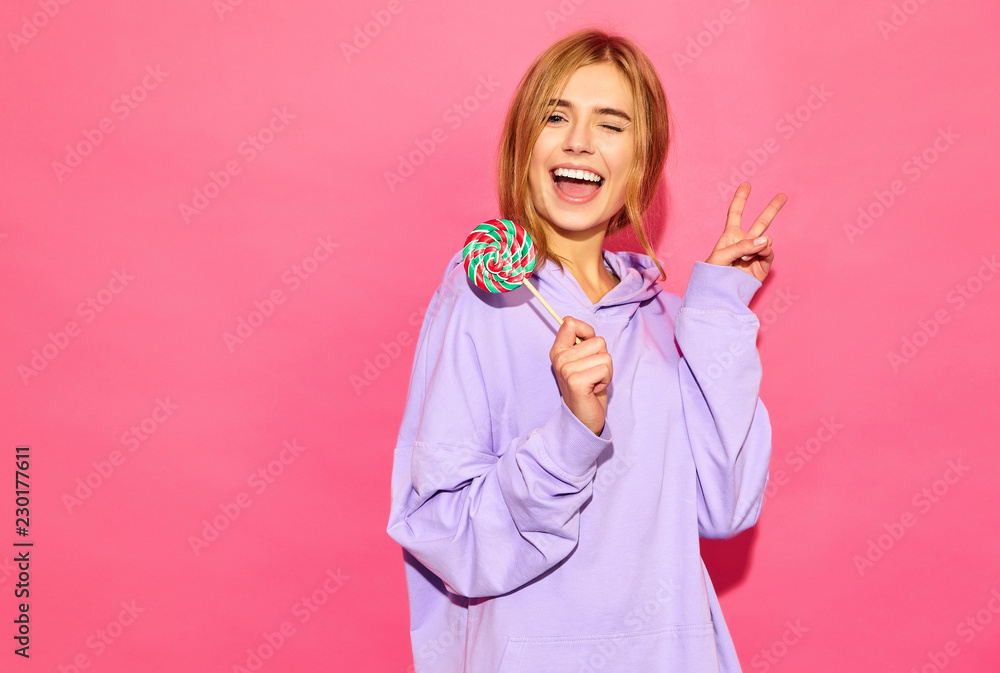 Portrait of young beautiful smiling hipster girl in trendy summer hoodie. Sexy carefree woman posing near pink wall. Positive model with lollipop showing peace sign. Portrait of young beautiful smiling hipster girl in trendy summer hoodie. Sexy carefree woman posing near pink wall. Positive model with lollipop showing peace sign.
