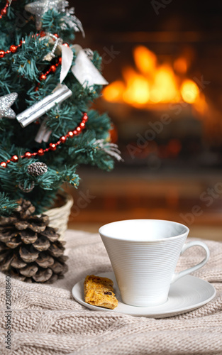 Cup of tea or coffee, woolen knitted things plaid and Christmas decorations near cozy fireplace background, in country house. Cozy relaxed magical atmosphere home interior. New Year winter concept.