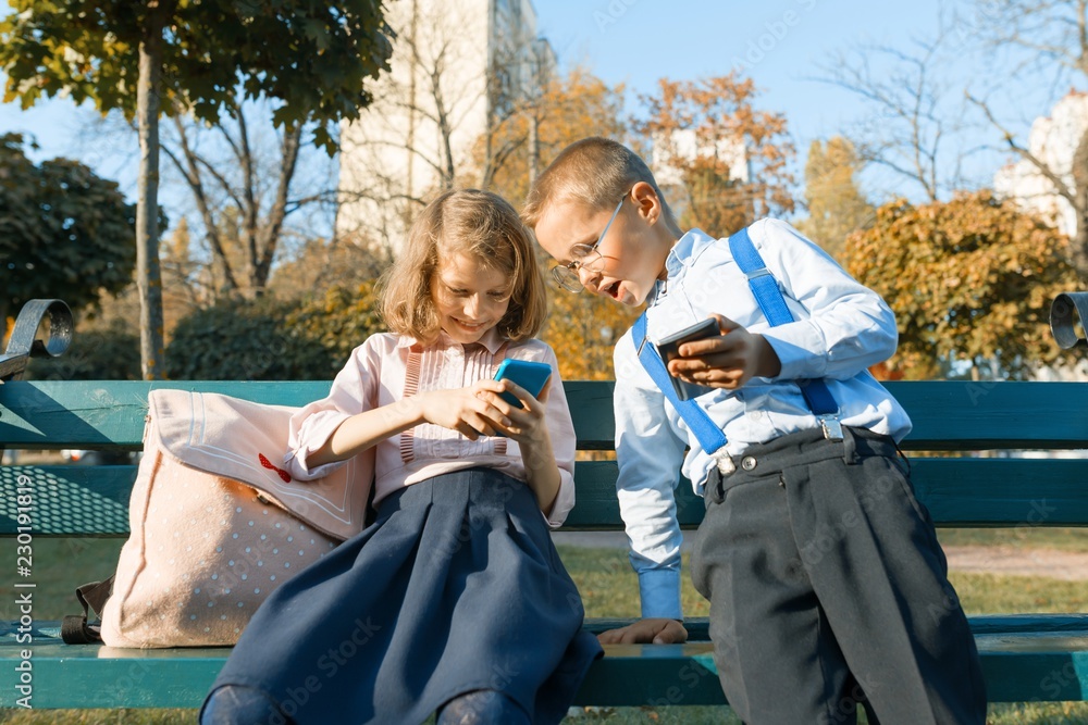 Fun children boy and girl are looking into smartphones. On a bench with school backpacks, background autumn sunny park, golden hour