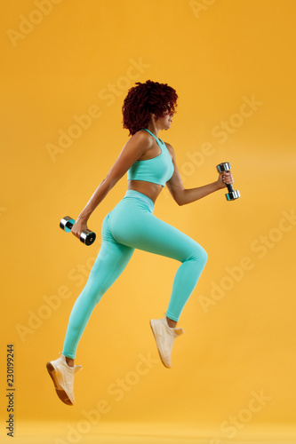 Strong athletic, woman sprinter or runner, running on yellow background with dumbbells wearing sportswear. Fitness and sport motivation.