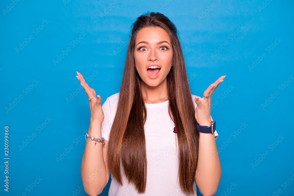 Fototapeta premium Cute brunette woman with long hair posing in white t-shirt on a blue background. Emotional portrait. She smiles in amazement