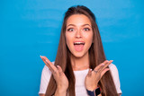 Cute brunette woman with long hair posing in white t-shirt on a blue background. Emotional portrait. She smiles in amazement