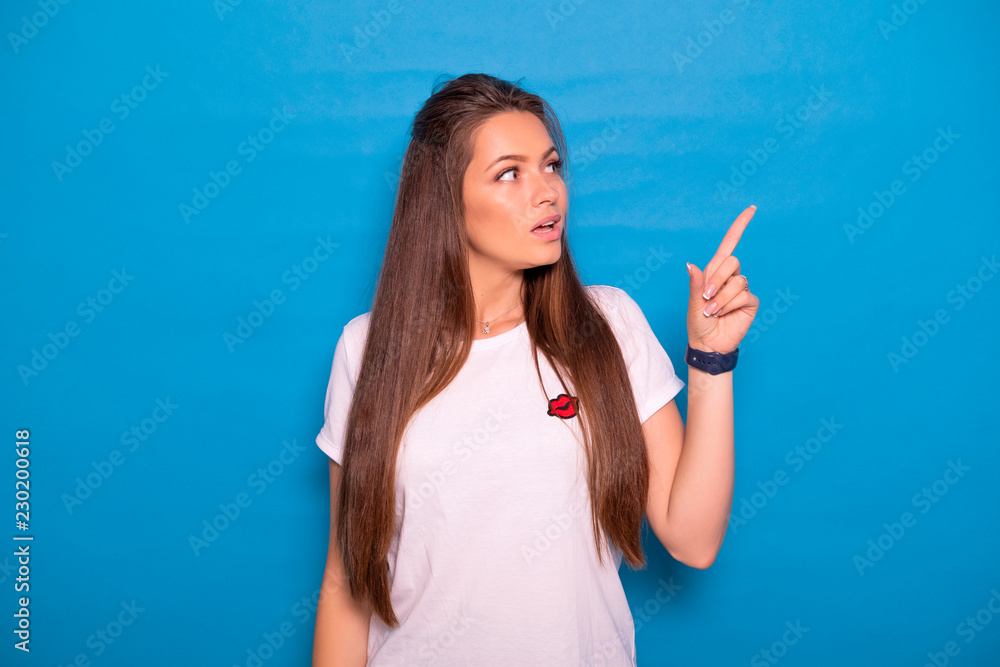 Fototapeta premium Cute brunette woman with long hair posing in white t-shirt on a blue background. Emotional portrait. She points her fingers on something, happily