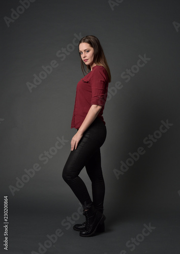 full length portrait of brunette girl wearing red shirt and leather pants. standing pose , on grey studio background.