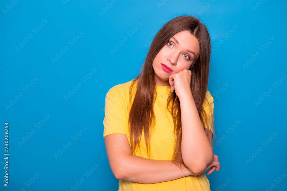 Cute brunette woman with long hair posing in yellow t-shirt on a blue background. Emotional portrait. She bored Cute brunette woman with long hair posing in yellow t-shirt on a blue background. Emotional portrait. She bored