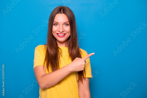 Cute brunette woman with long hair posing in yellow t-shirt on a blue background. Emotional portrait. She smiles happily with flawless white teeth and poins finger on something