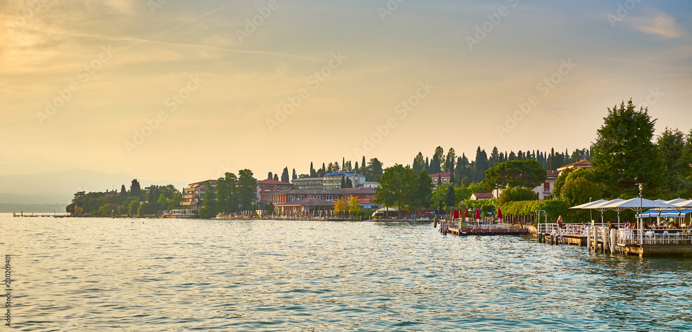 City of Sirmione at Lake Garda at sunset / People sitting in a restaurant next to harbor with boats 