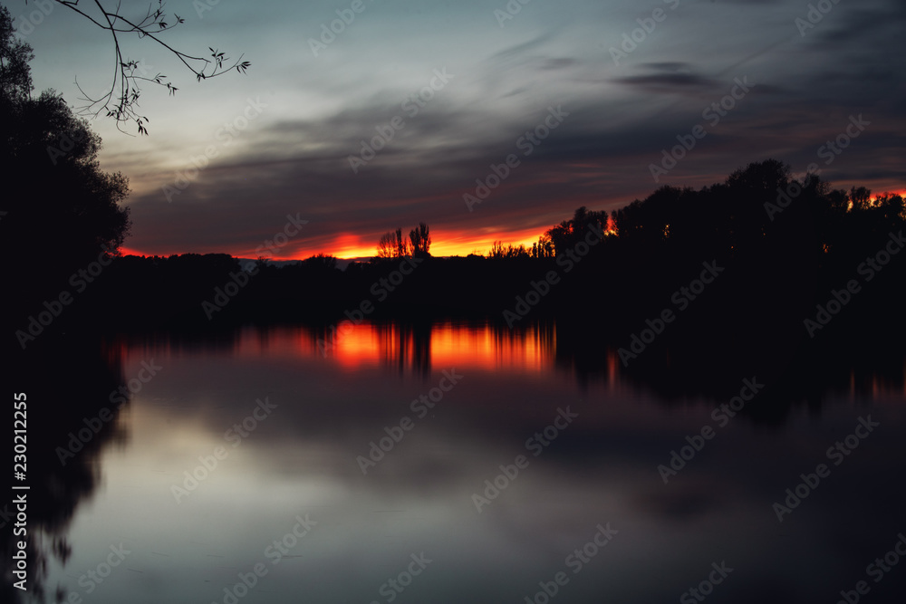 Dramatic long exposure of a calm lake with blurry clouds and sunset light and tree reflection. Südsee in Braunschweig, Germany
