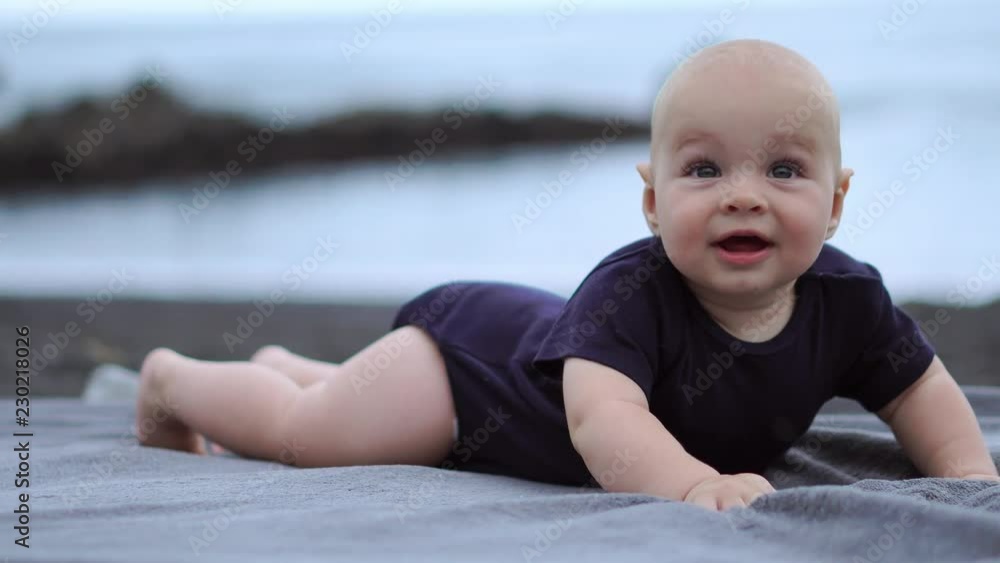 The baby lies on his stomach on the black sand near the ocean and laughs looking at the camera The baby lies on his stomach on the black sand near the ocean and laughs looking at the camera