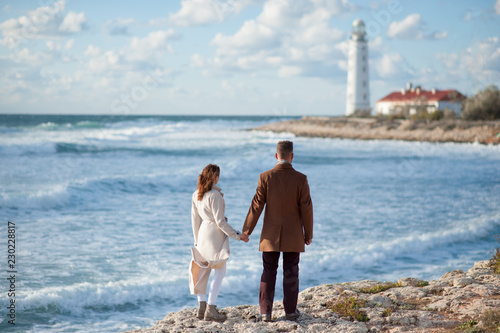 young love couple consisting of man and woman in coats standing near stormy sea coast with lighthouse