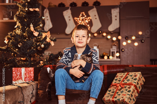 Adorable boy hugging his cat in hands while sitting on sofa in decorated room at Christmas time.