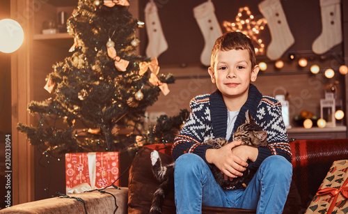 Adorable boy hugging his cat in hands while sitting on sofa in decorated room at Christmas time.